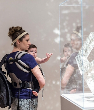 A mother carries a child, who points at an encased sculptural artwork